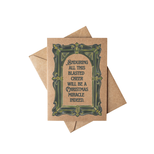 A square kraft paper greeting card with a decorative dark green ornate border and humorous text reading 'ENDURING ALL THIS BLASTED CHEER WILL BE A CHRISTMAS MIRACLE INDEED.' A matching kraft envelope is placed behind it on a clean white background.