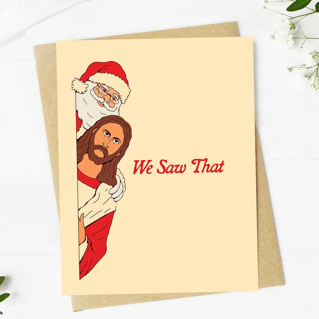 A cream-colored greeting card with a brown kraft envelope, featuring a cartoon illustration of Jesus Christ and Santa Claus peeking from behind a wall with the text 'We Saw That' in red. The card is displayed on a white wooden surface with small green leaves and white flowers in the corners.