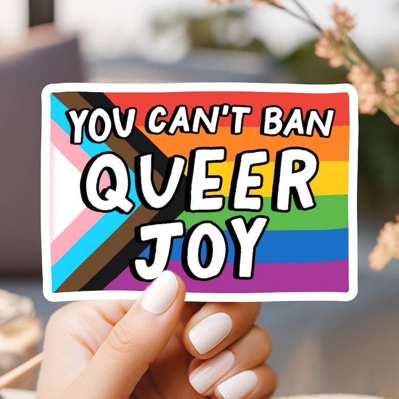 Hand holding a rainbow-colored sticker with text 'YOU CAN'T BAN QUEER JOY' against a blurred background.