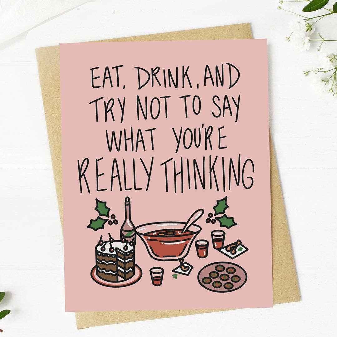A pink greeting card with a humorous quote and a holiday-themed illustration, displayed on top of a brown kraft envelope against a white wooden background.