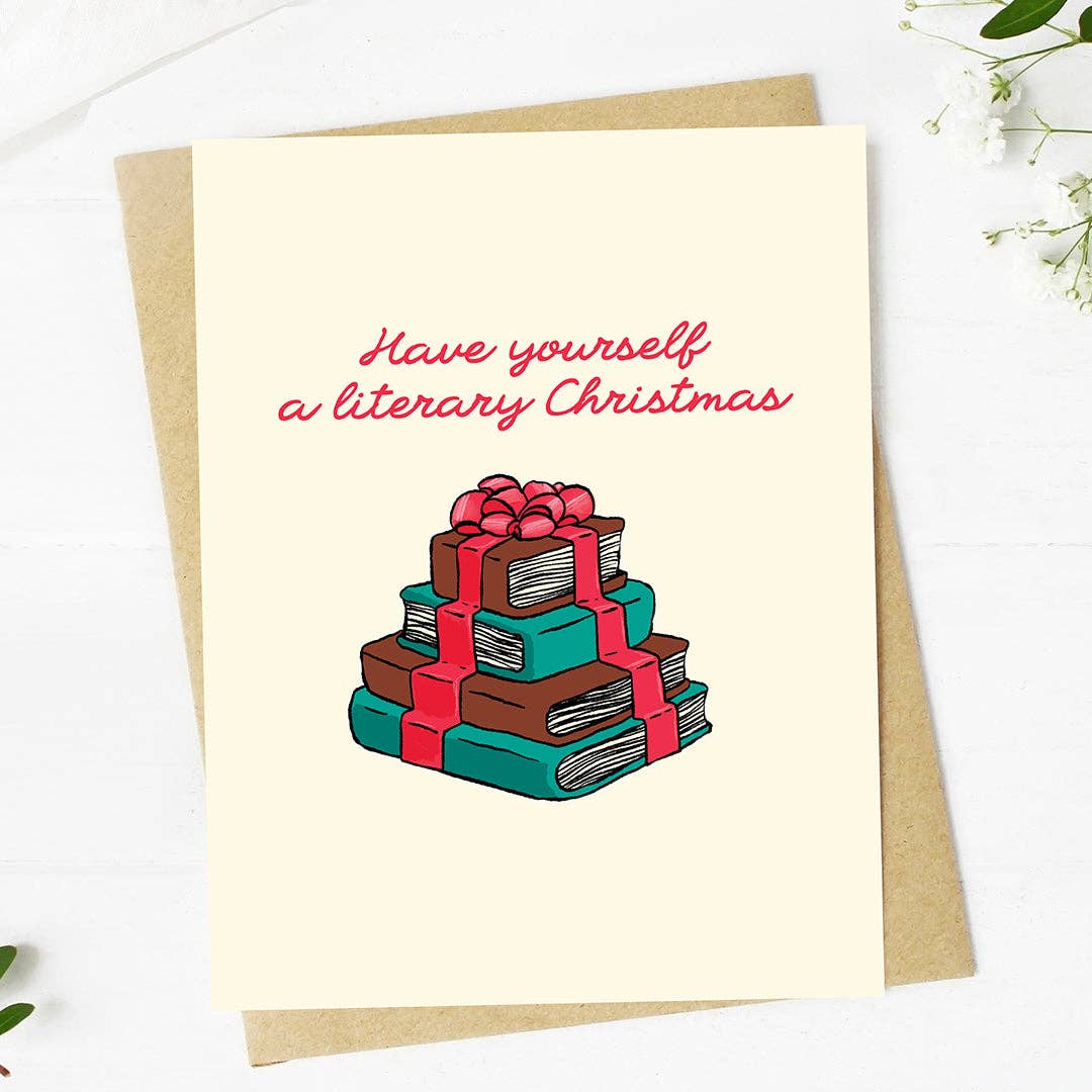 A cream-colored greeting card with the text 'Have yourself a literary Christmas' in pink script above an illustration of a stack of books wrapped in red ribbon, shown with a brown kraft envelope on a white background.