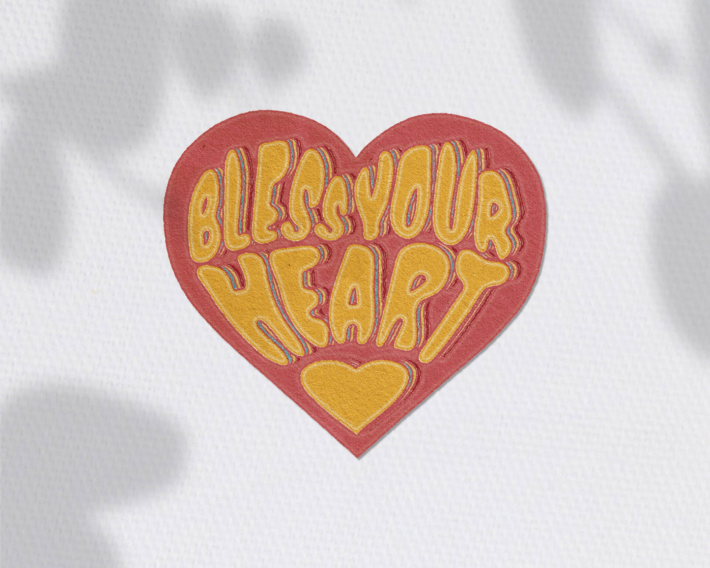 A heart-shaped patch with a red border and yellow text reading 'Bless Your Heart' on a white textured background with soft leaf shadows.