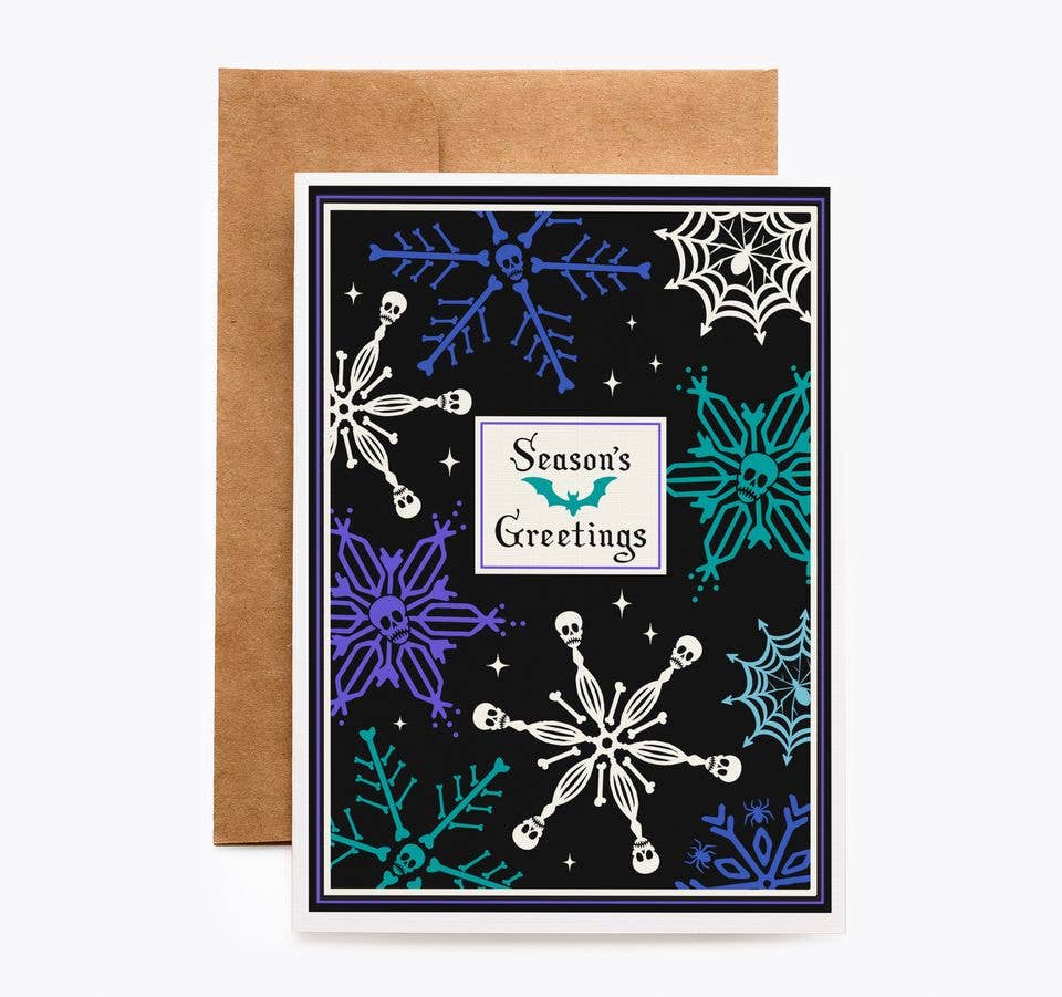A Halloween-themed greeting card with a brown kraft envelope behind it. The card has a black background decorated with white, blue, and teal snowflakes, spiderwebs, stars, and a central white label that reads 'Season's Greetings'.