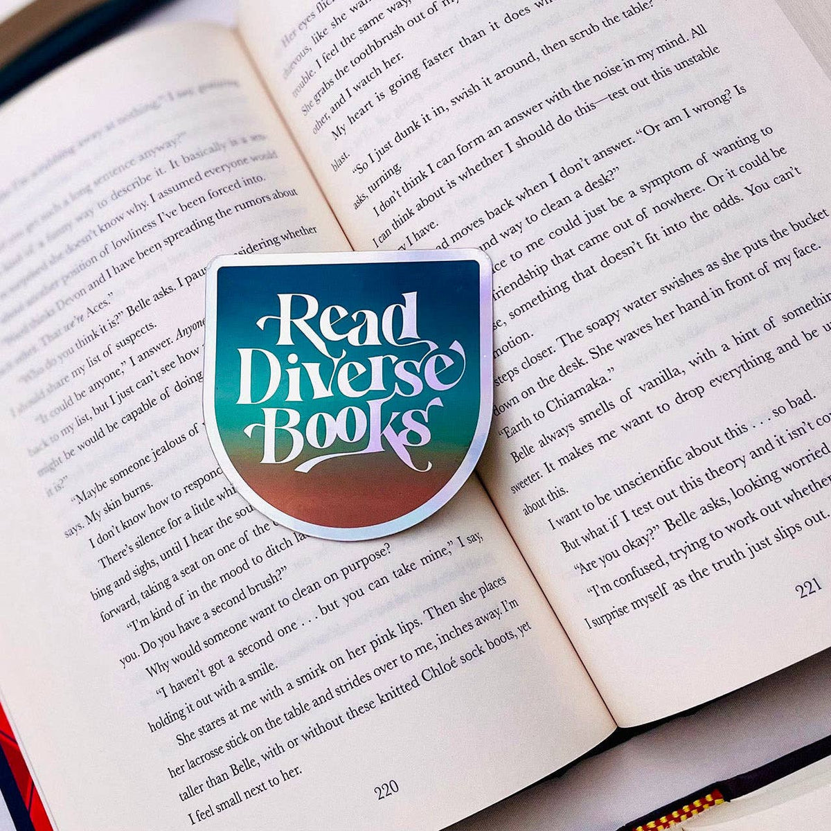 A shield-shaped book bookmark with the words 'Read Diverse Books' in white script over a teal-to-brown gradient, placed on open book pages.
