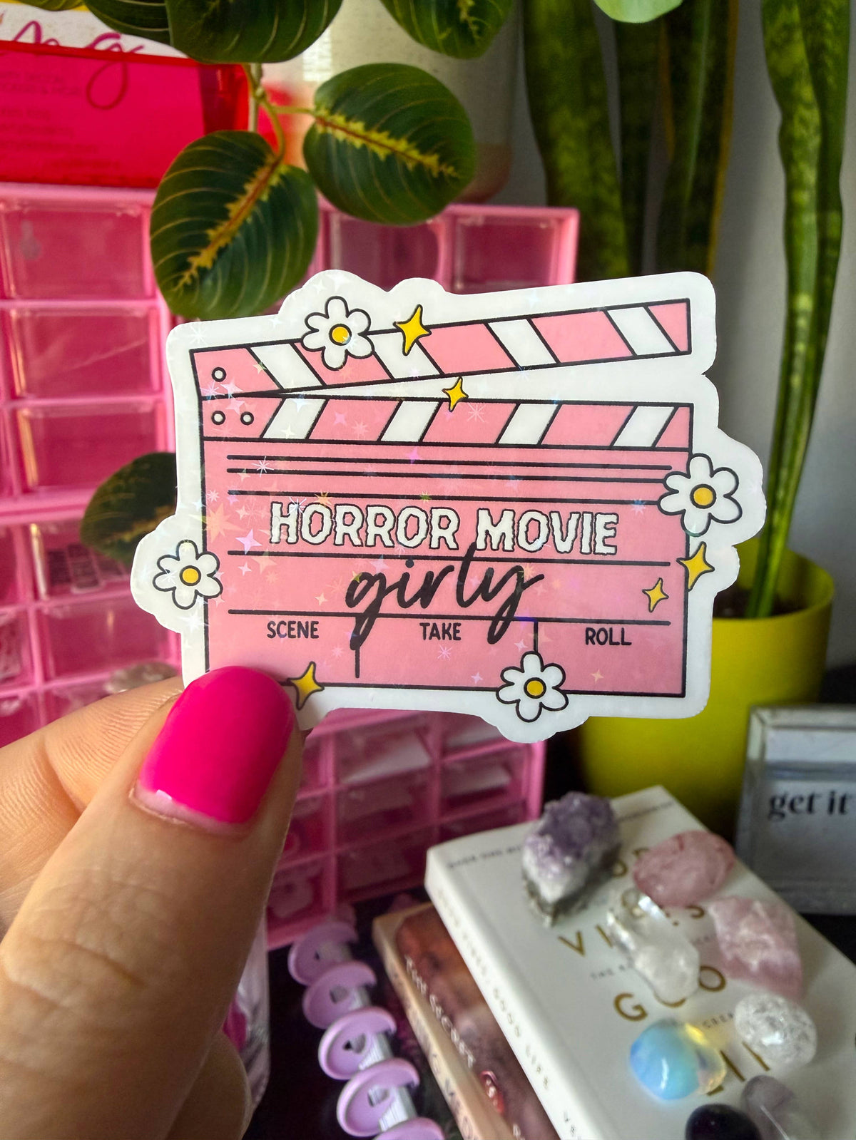 A hand with bright pink nail polish holds a die-cut sticker shaped like a pink horror movie clapperboard. The sticker has white daisy flowers, yellow stars, and the text 'HORROR MOVIE girly' with 'SCENE TAKE ROLL' underneath.