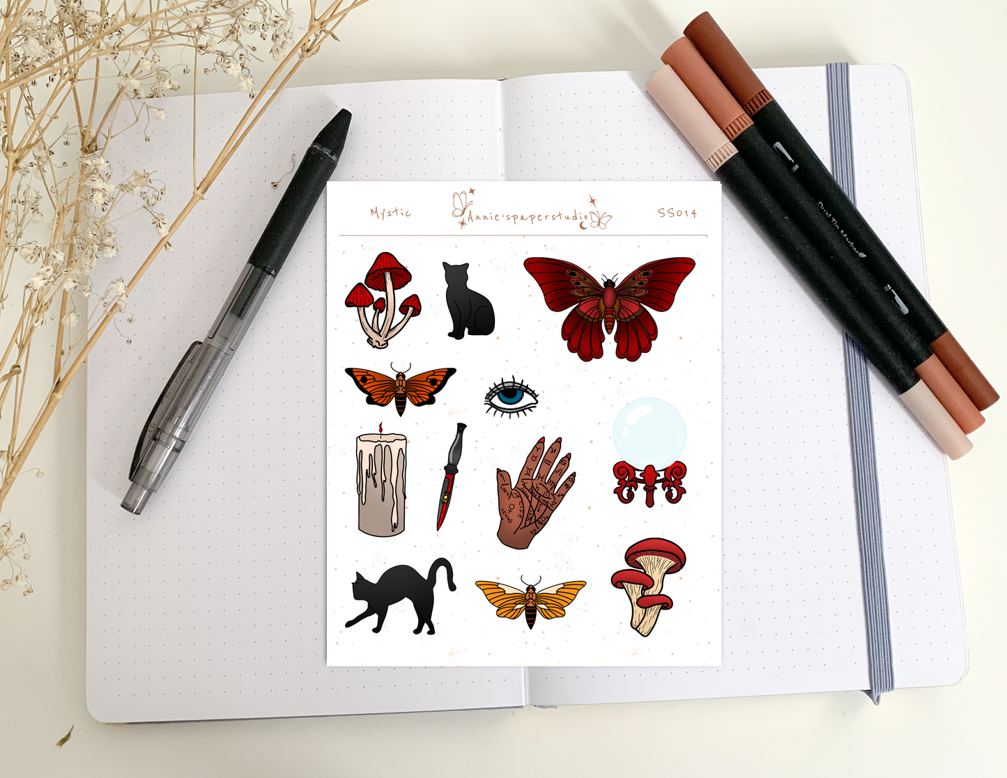 A sheet of decorative stickers displayed on an open notebook, surrounded by a black pen, two brown makeup brushes, and dried flowers. The sticker sheet features whimsical illustrations including red and orange butterflies, black cats, red mushrooms, a candle, a dagger, a hand, and an eye.