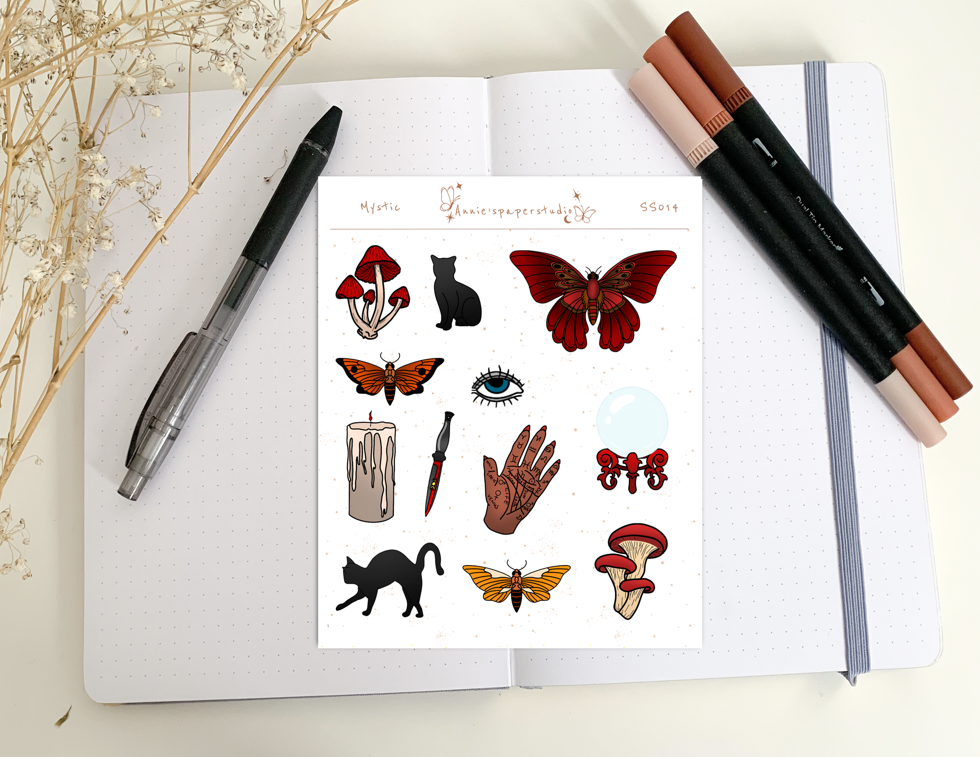 A sheet of decorative stickers displayed on an open notebook, surrounded by a black pen, two brown makeup brushes, and dried flowers. The sticker sheet features whimsical illustrations including red and orange butterflies, black cats, red mushrooms, a candle, a dagger, a hand, and an eye.