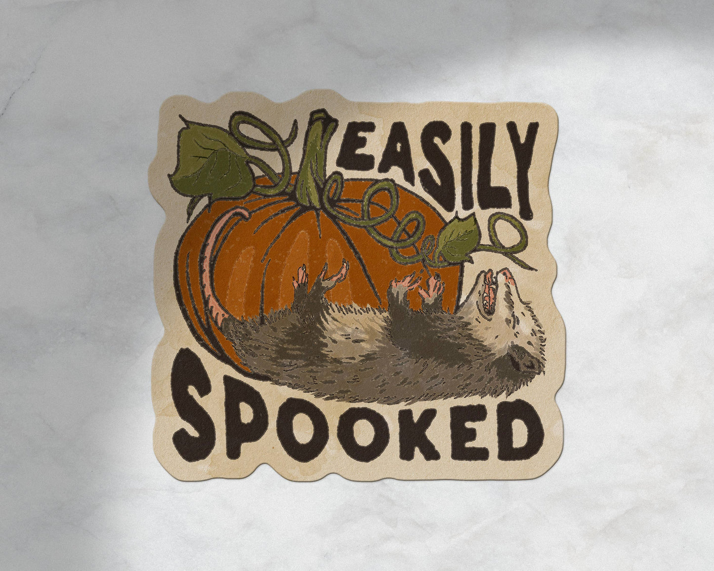 A die-cut sticker featuring a cartoon pumpkin and hedgehog with the text 'EASILY SPOOKED' on a light marble background.