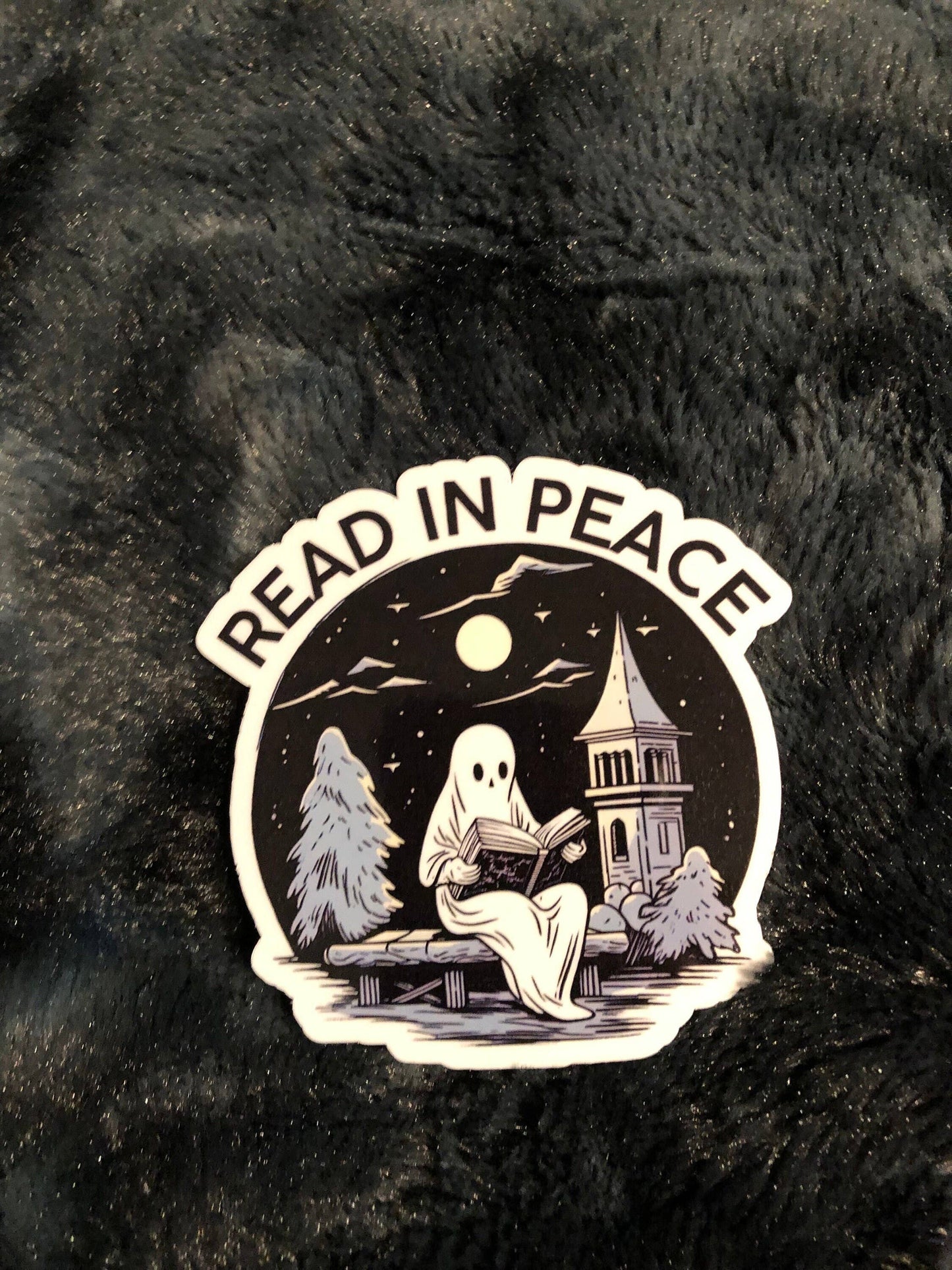A die-cut sticker featuring a spooky night scene with a ghost reading on a bench under a full moon, with the words 'READ IN PEACE' arched above, placed on a dark fuzzy fabric background.