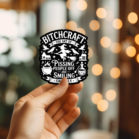 Hand holding a sticker with 'Bitchcraft' design against a blurred background with lights.