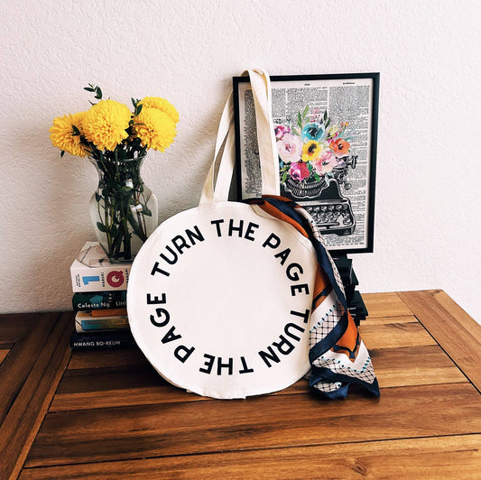Tote bag with 'Turn the Page' text on a wooden surface with a vase of flowers and framed art in the background.