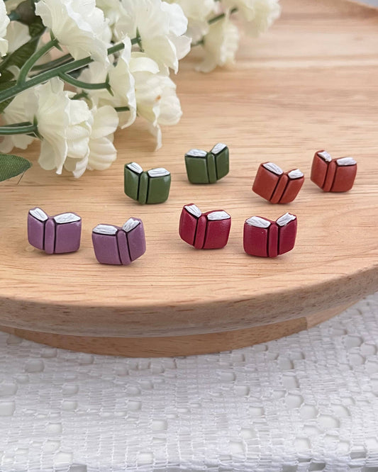 Set of small book-shaped earrings on a wooden surface with white flowers in the background.