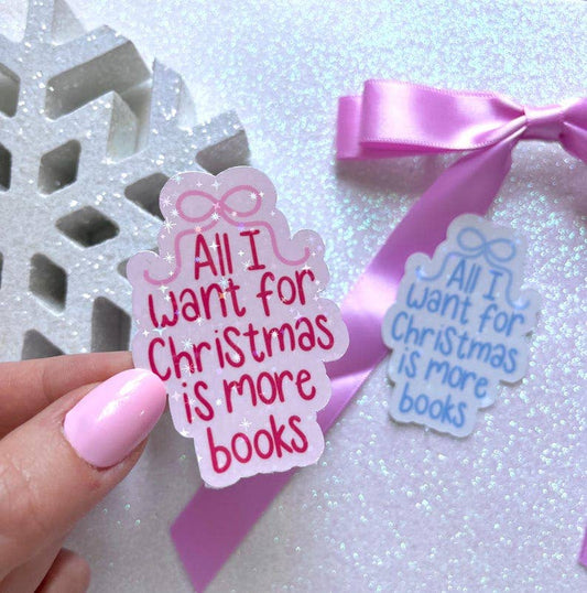A hand with pink nail polish holds a scalloped gift box-shaped sticker that reads 'All I want for Christmas is more books' in pink lettering on a white background. A second matching sticker with blue lettering lies nearby on a glittery white surface next to a pink ribbon bow.