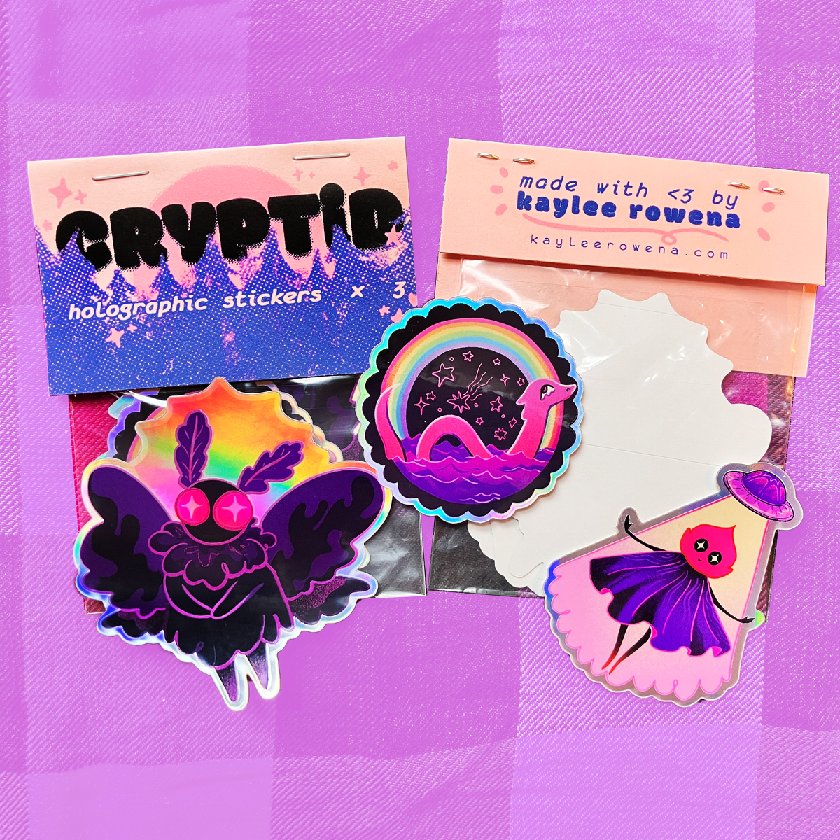 A set of colorful holographic stickers displayed on a purple plaid background. The stickers include a purple winged creature with pink eyes, a rainbow with a pink snake, and a slice of cake with a face, alongside branded backing cards reading 'CRYPTID' and 'made with <3 by kaylee rowana'.