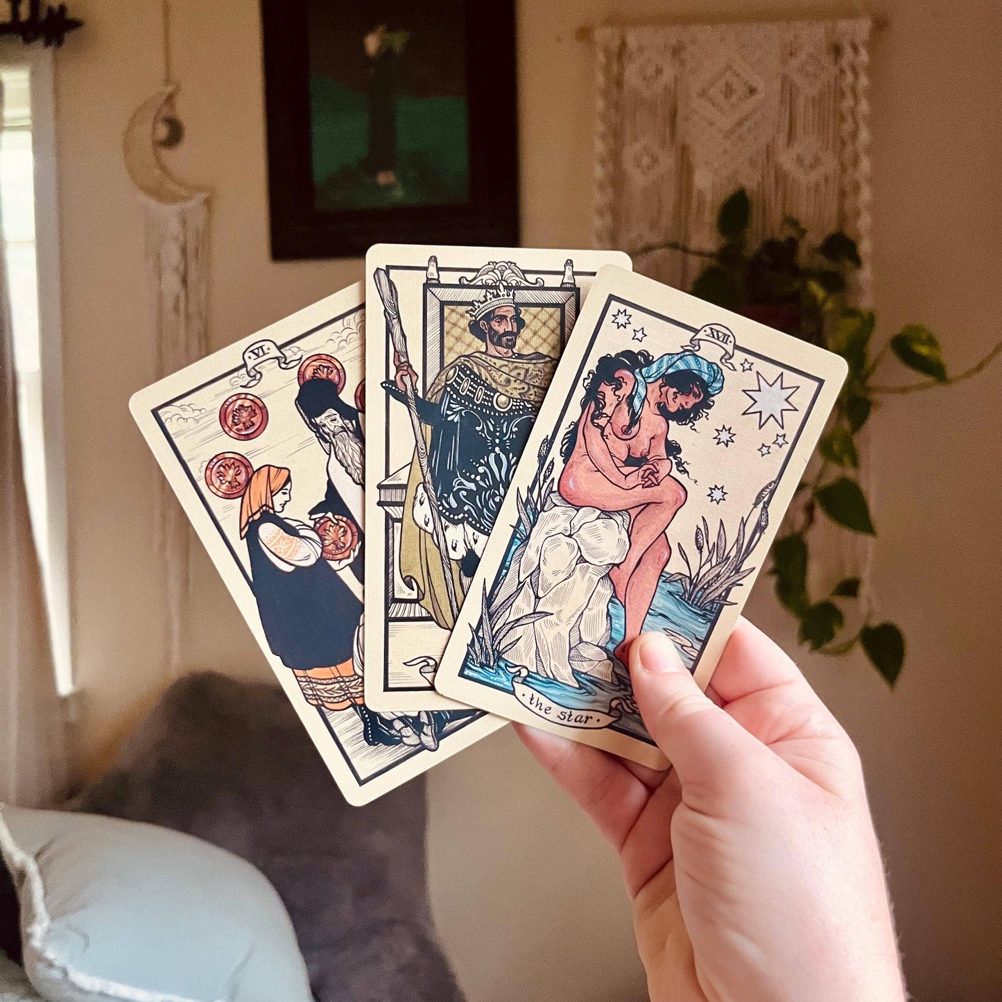 Three tarot cards held by a hand in a cozy room with decor.