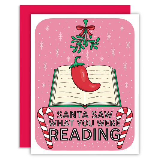 A Christmas-themed greeting card with a pink background and red envelope. The card features an illustration of an open book with a red chili pepper on it, mistletoe, candy canes, and the text 'SANTA SAW WHAT YOU WERE READING'.