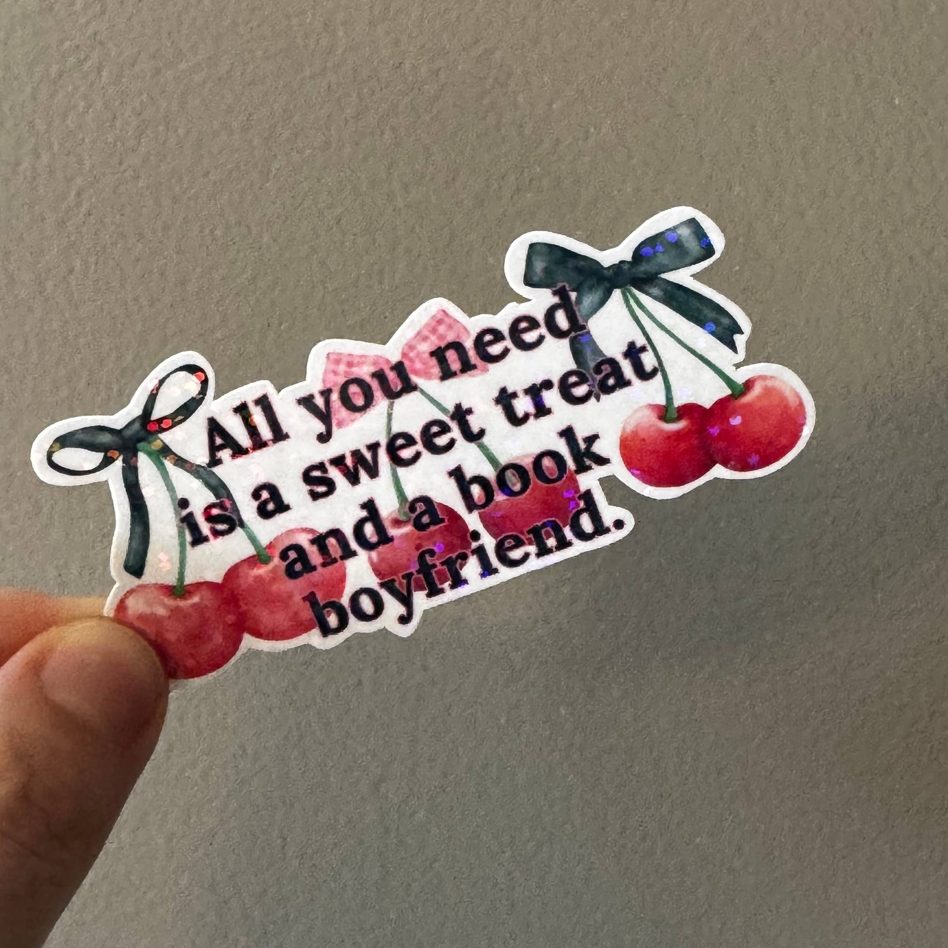 A hand holding a die-cut sticker with the quote 'All you need is a sweet treat and a book boyfriend.' in black text, decorated with red cherries, pink hearts, and black bows, against a plain gray wall.