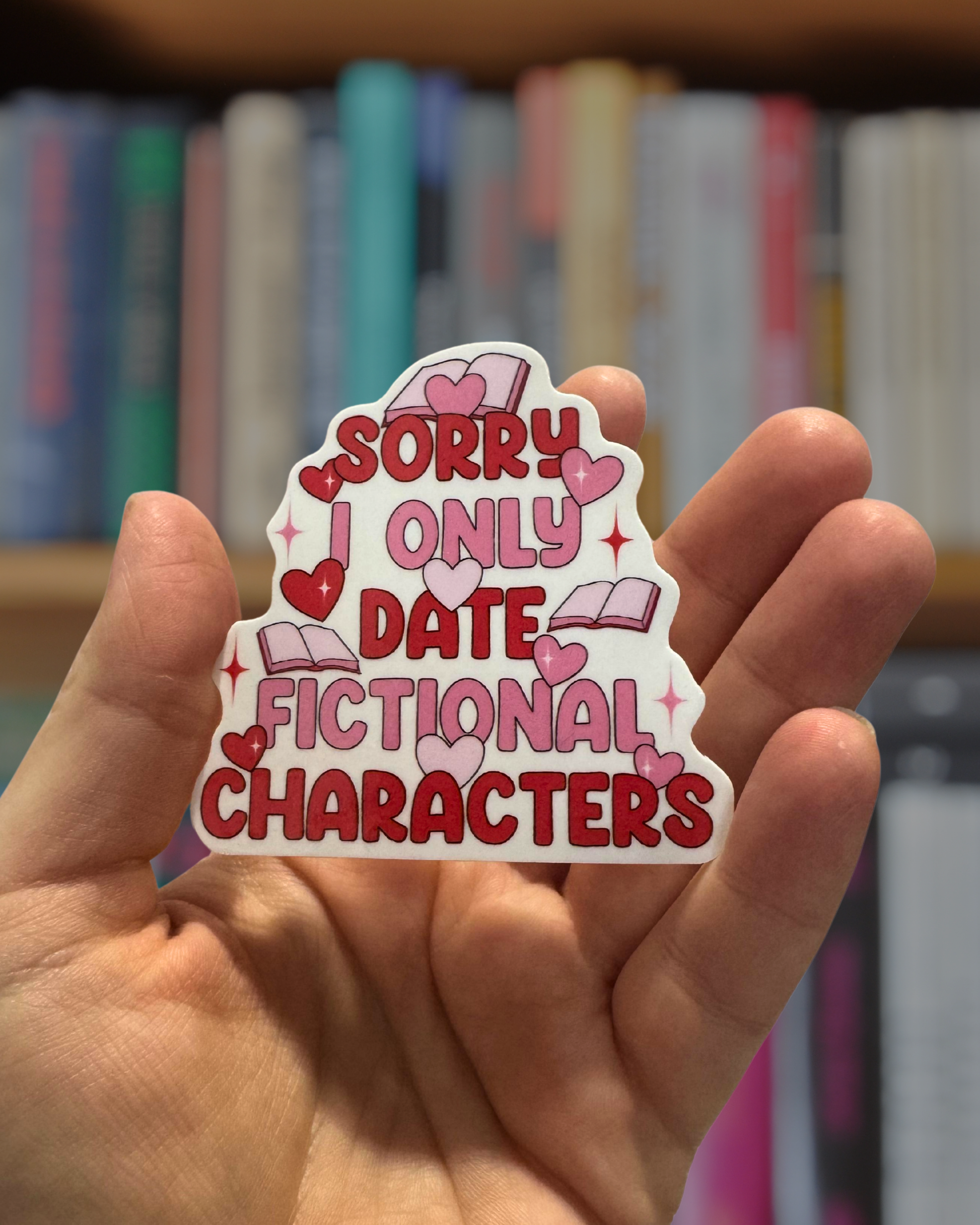 Hand holding a sticker with text 'Sorry I only date fictional characters' against a bookshelf background