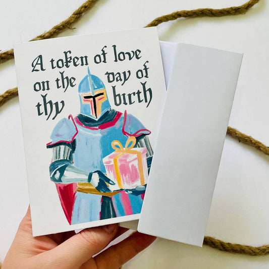 Card with a character holding a gift and text 'A token of love on the day of thy birth' held by a hand.