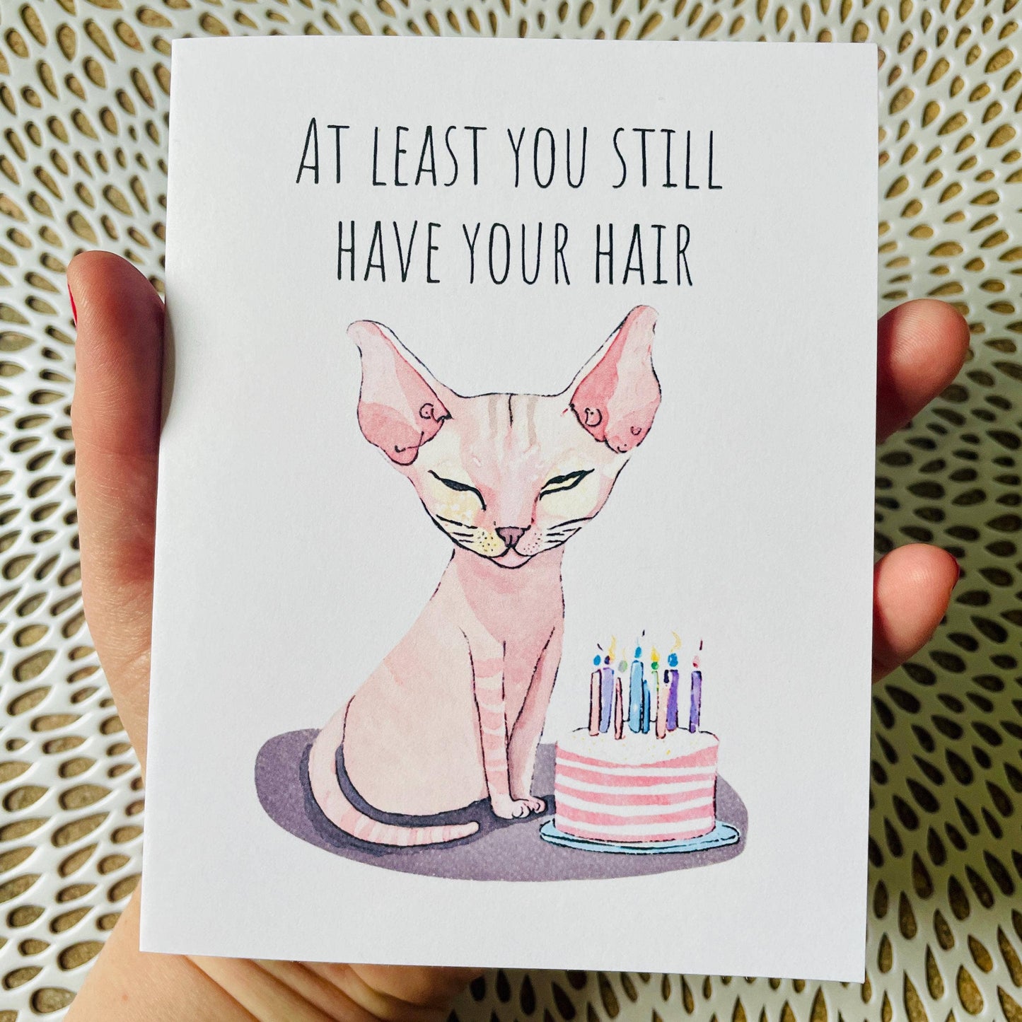 Card with a cat and birthday cake illustration and text 'At least you still have your hair' held by a hand.