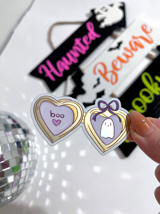 A hand holds a pair of novelty heart-shaped sunglasses with a gold frame and purple lenses, one lens showing the word 'boo' and the other a ghost illustration. The background is a white surface with Halloween-themed signs and a disco ball.