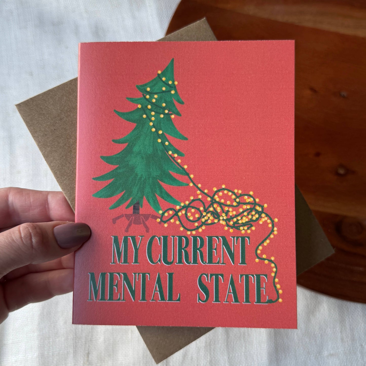 A hand holds a coral-pink greeting card featuring a green Christmas tree with tangled yellow string lights and the text 'MY CURRENT MENTAL STATE' in teal. A brown kraft envelope is partially visible behind the card, with a white fabric surface and a wooden surface in the background.