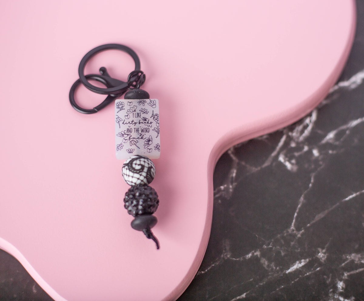 A black metal keychain with a rectangular white bead printed with black floral patterns, a black spiral bead, and a black textured seed bead, displayed on a pink guitar-shaped board over a dark marble surface.