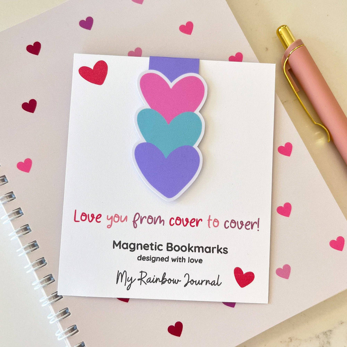 Magnetic bookmarks with heart designs on a card labeled 'My Rainbow Journal' with a pen and notebook in the background.