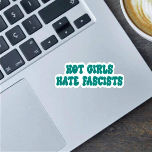Laptop with a sticker reading 'Hot Girls Hate Fascists' next to a cup of coffee.