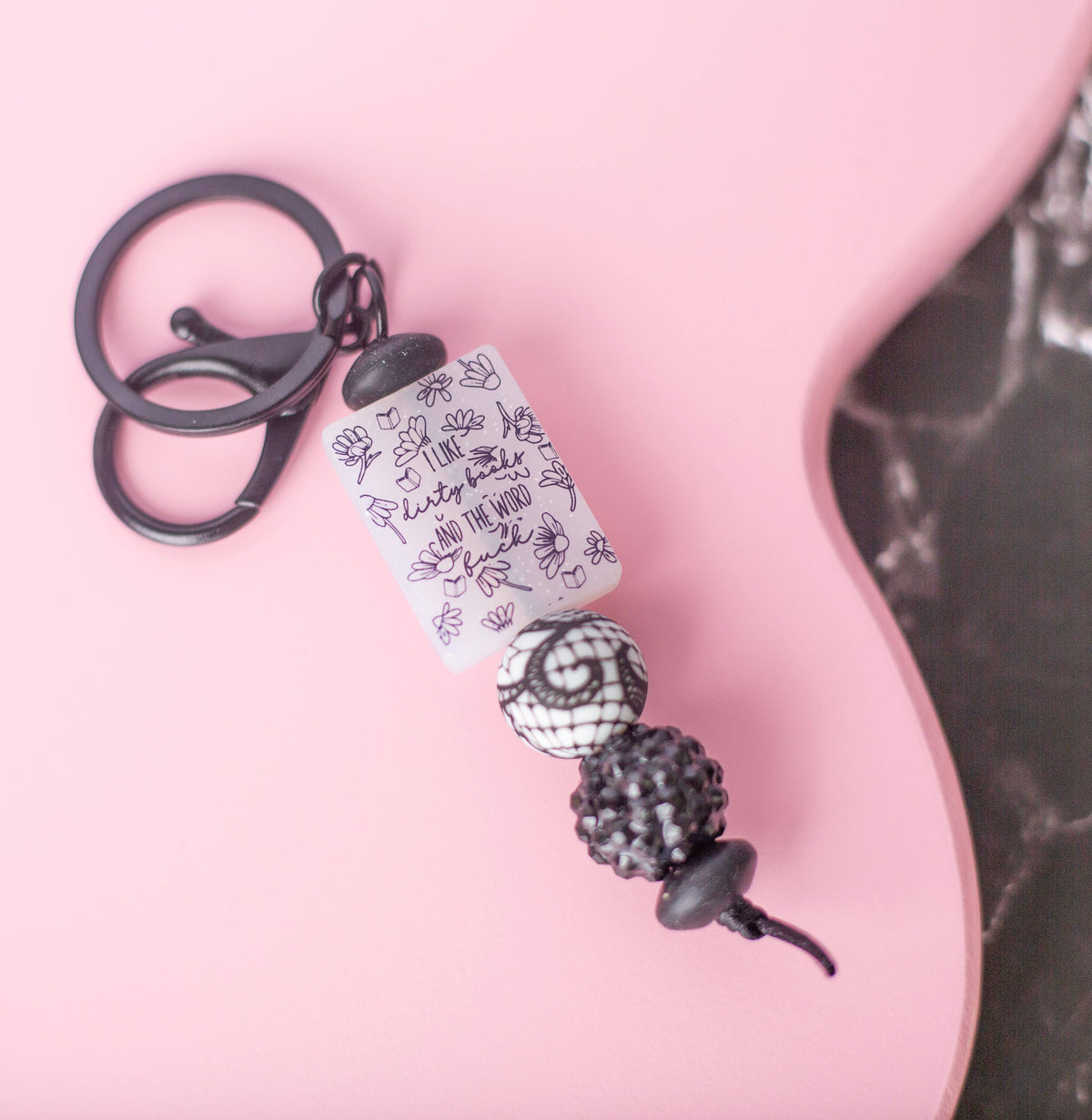 A black metal keychain with a clear rectangular charm printed with black hand-drawn illustrations and text, plus decorative black and white beads, styled on a pink background.