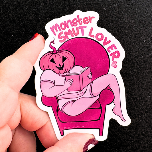 Sticker with a pink pumpkin character reading a book and 'Monster SMUT LOVER' text, held by a hand against a black background.