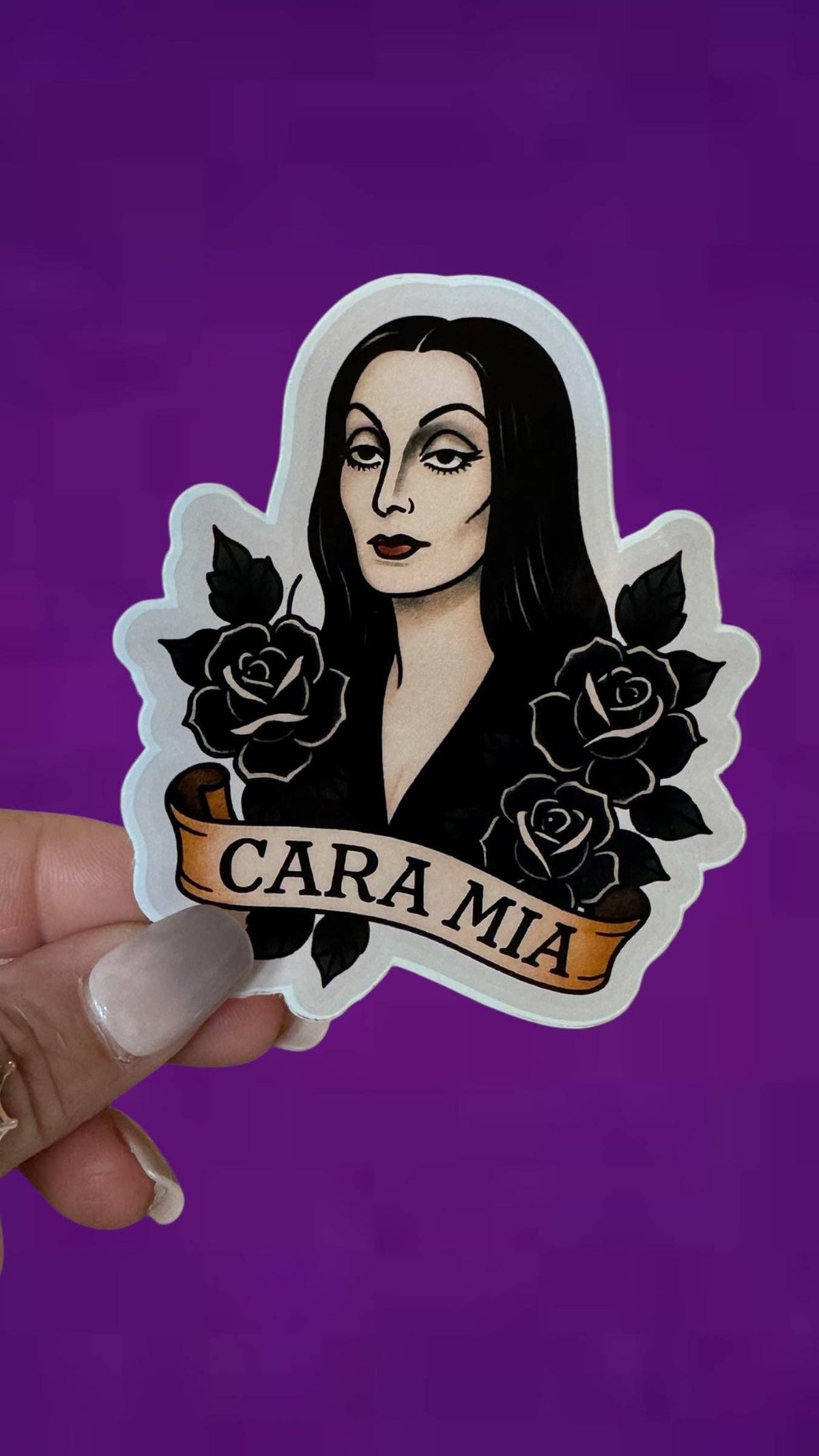 A hand holds a die-cut sticker featuring an illustrated woman with long black hair, black roses, and a banner reading 'CARA MIA' against a solid purple background.