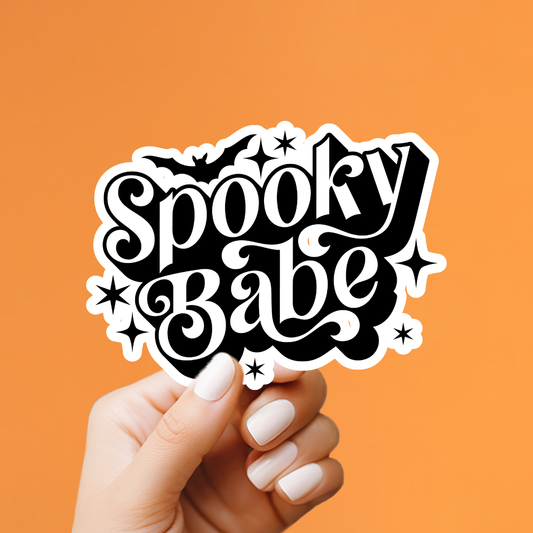 A hand with white manicured nails holds a die-cut sticker that reads 'Spooky Babe' in black retro-style lettering with white stars and bats, set against a solid orange background.