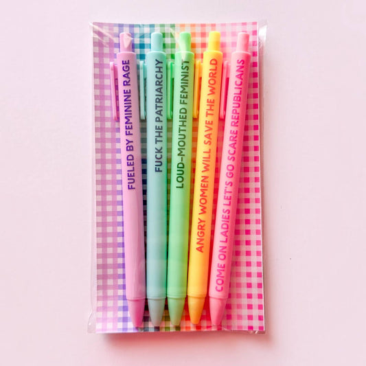 A set of five novelty highlighter pens in pink, teal, green, yellow, and orange, packaged in a clear plastic sleeve over a pink and white gingham patterned backing card. The pens feature bold, humorous feminist slogans printed vertically on their barrels.