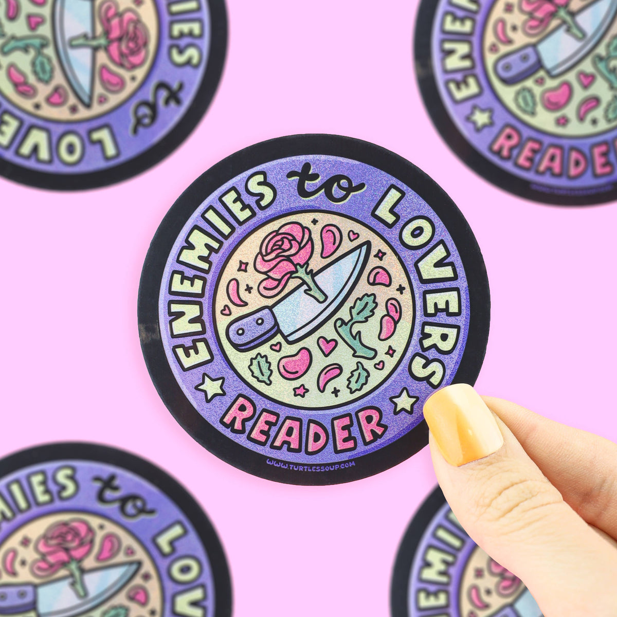 Sticker with 'Enemies to Lovers Reader' design held by a hand on a pink background