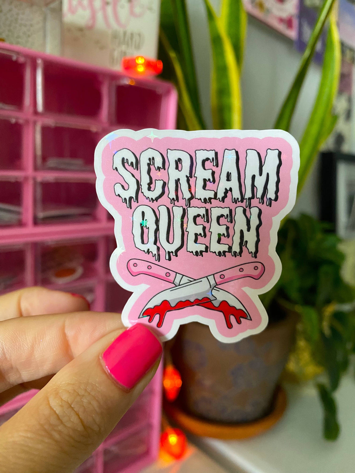 A hand with bright pink nail polish holds a die-cut sticker that reads 'SCREAM QUEEN' in dripping white letters on a pink background with crossed knives and blood splatter. A pink organizer and potted plants are blurred in the background.