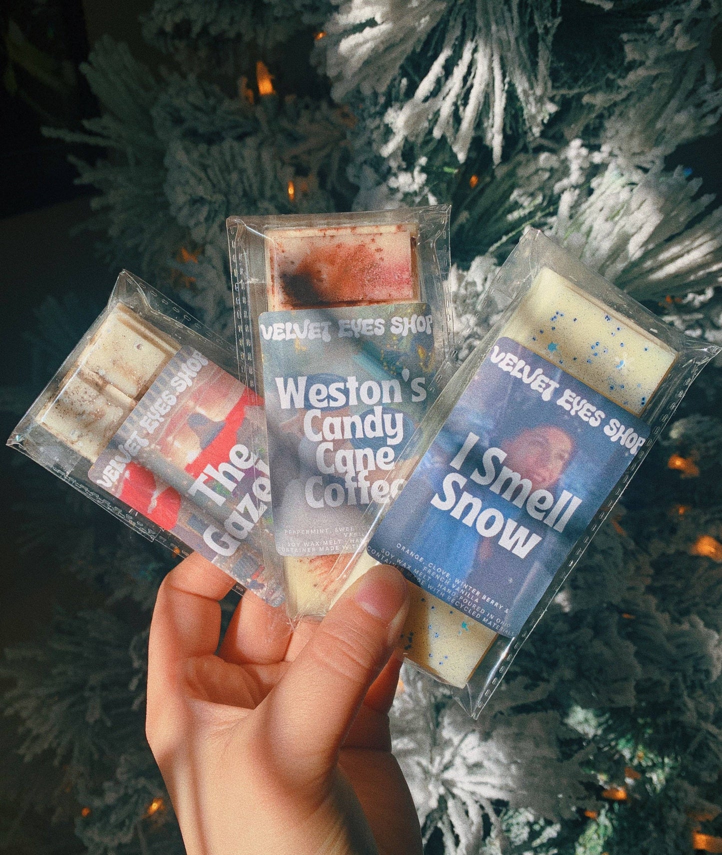 Three scented candles in packaging held in front of a decorated Christmas tree.