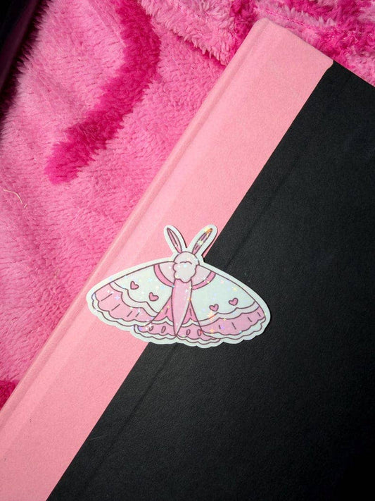 A close-up of a notebook or journal with a diagonal split design in pink and black, featuring a die-cut sticker of a bunny in a pink dress on the pink section. The background shows a fuzzy hot pink blanket.