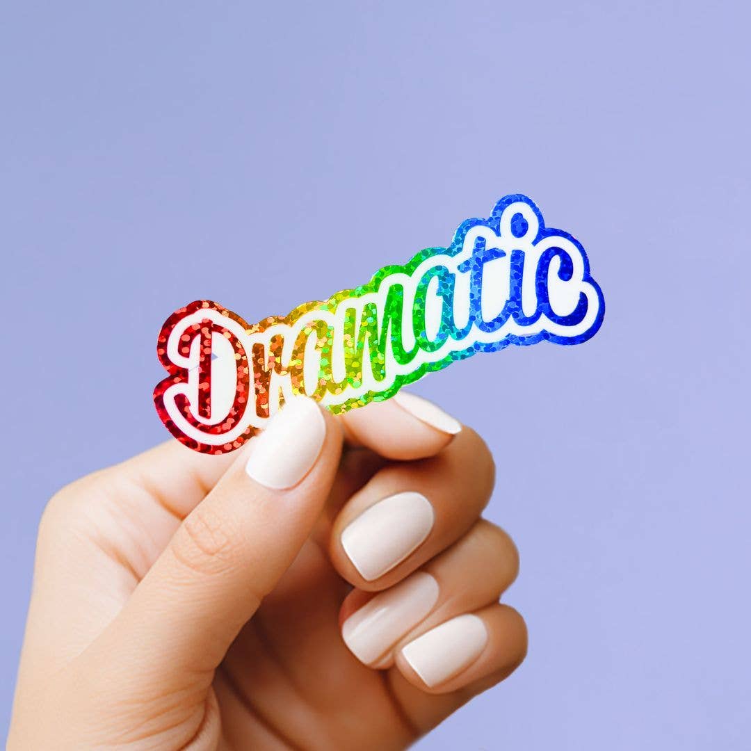 Hand holding a colorful 'Dramatic' sticker against a purple background