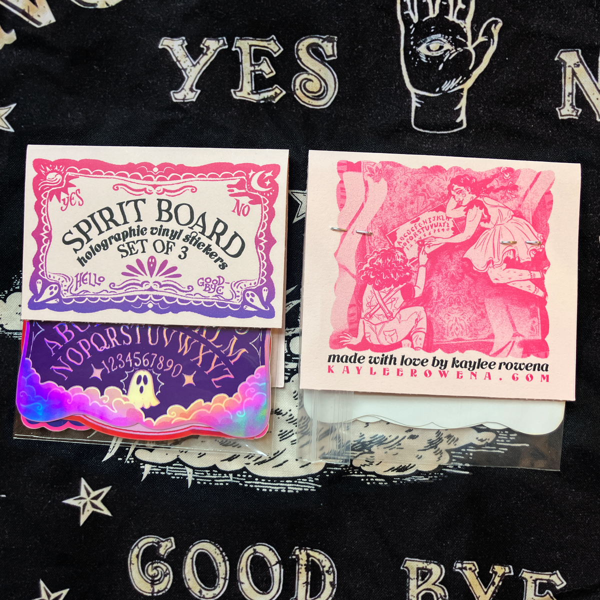 A set of three occult-themed holographic vinyl stickers displayed on a black fabric background. The stickers feature gothic designs including a 'Spirit Board' Ouija board illustration and a pink-toned illustration of a woman kneeling before a board, with one sticker showing a colorful holographic rainbow border.