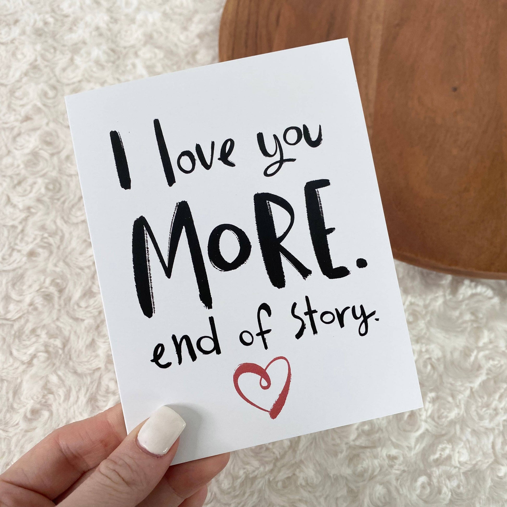 Hand holding a card with 'I love you MORE. end of story.' text and a red heart on a textured surface.