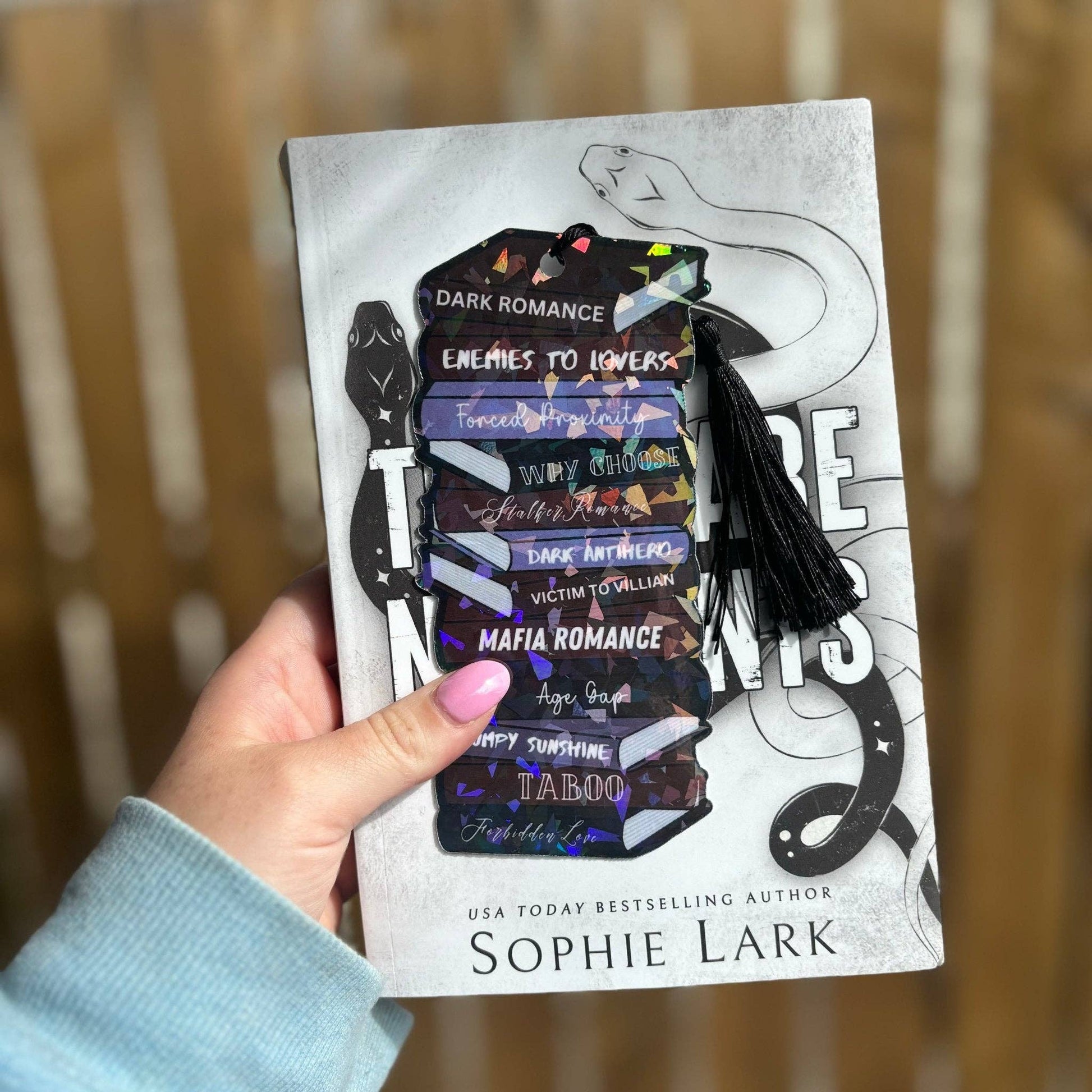 A hand holding a cardstock bookmark with 'Dark Romance' and a list of romance trope names printed on it, with a black and white book cover in the background.