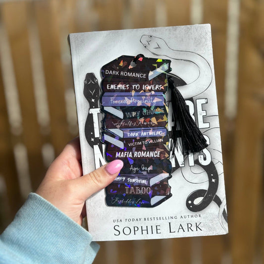 A hand holding a cardstock bookmark with 'Dark Romance' and a list of romance trope names printed on it, with a black and white book cover in the background.
