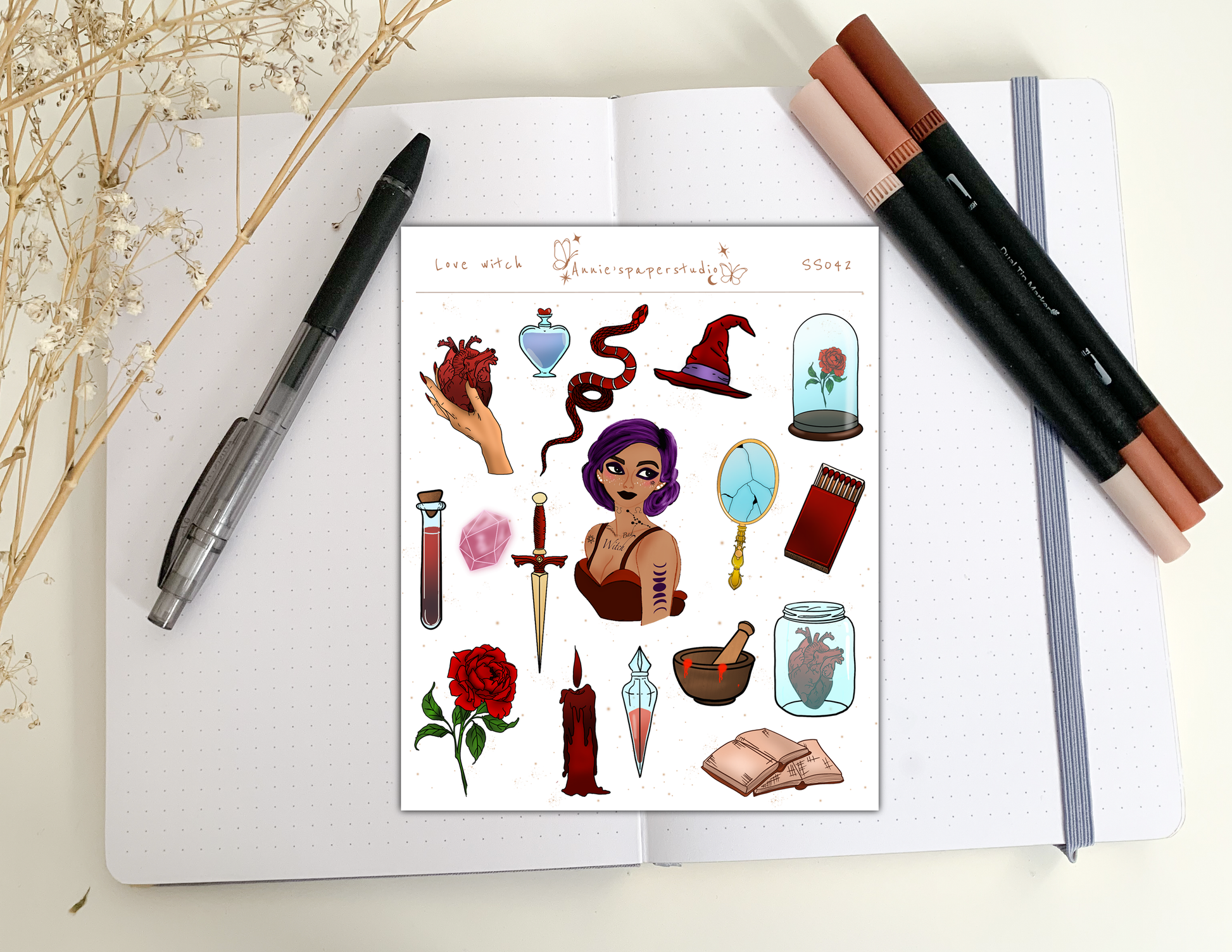 A sheet of illustrated stickers featuring gothic and witch-themed designs, displayed on an open notebook with decorative pens and dried flowers.
