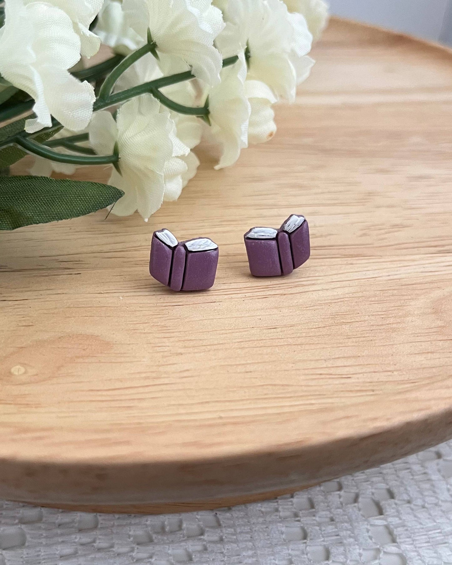 Purple book-shaped earrings on a wooden surface with white flowers in the background