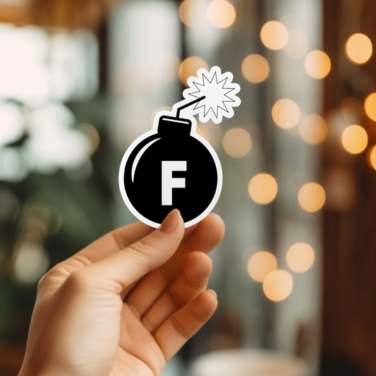 Hand holding a bomb-shaped sticker with an 'F' on a blurred background