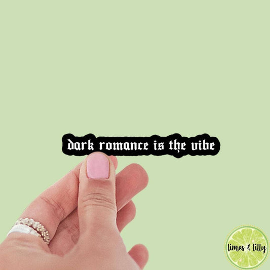 Hand with pink nail polish holding a black sign with 'dark romance is the vibe' text on a light green background.