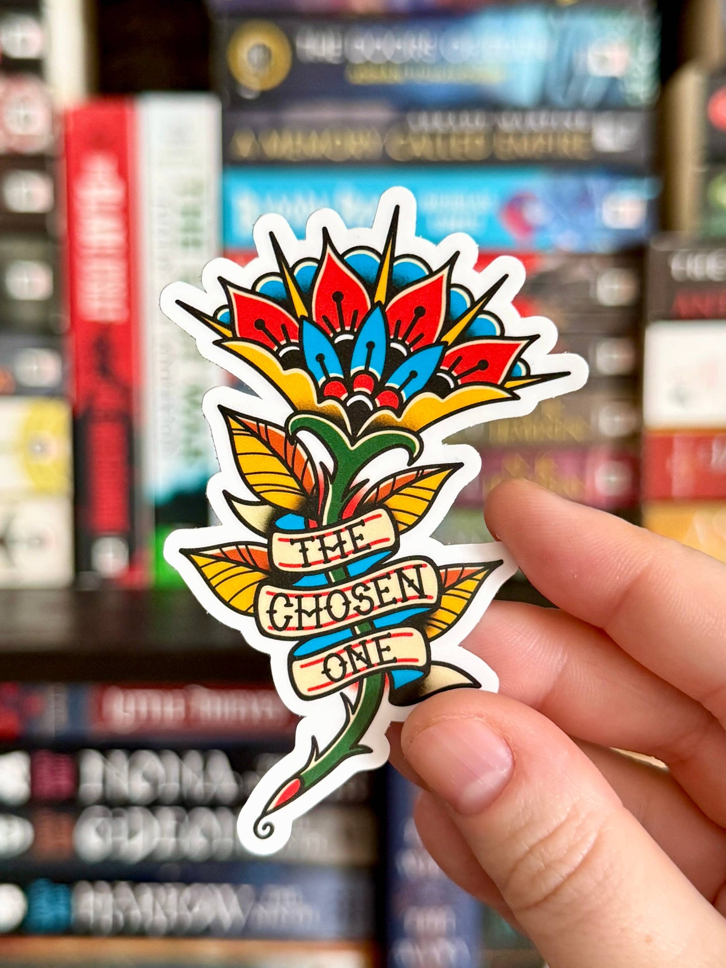 Colorful tattoo-style sticker with 'The Chosen One' text held by a hand against a blurred bookshelf background.