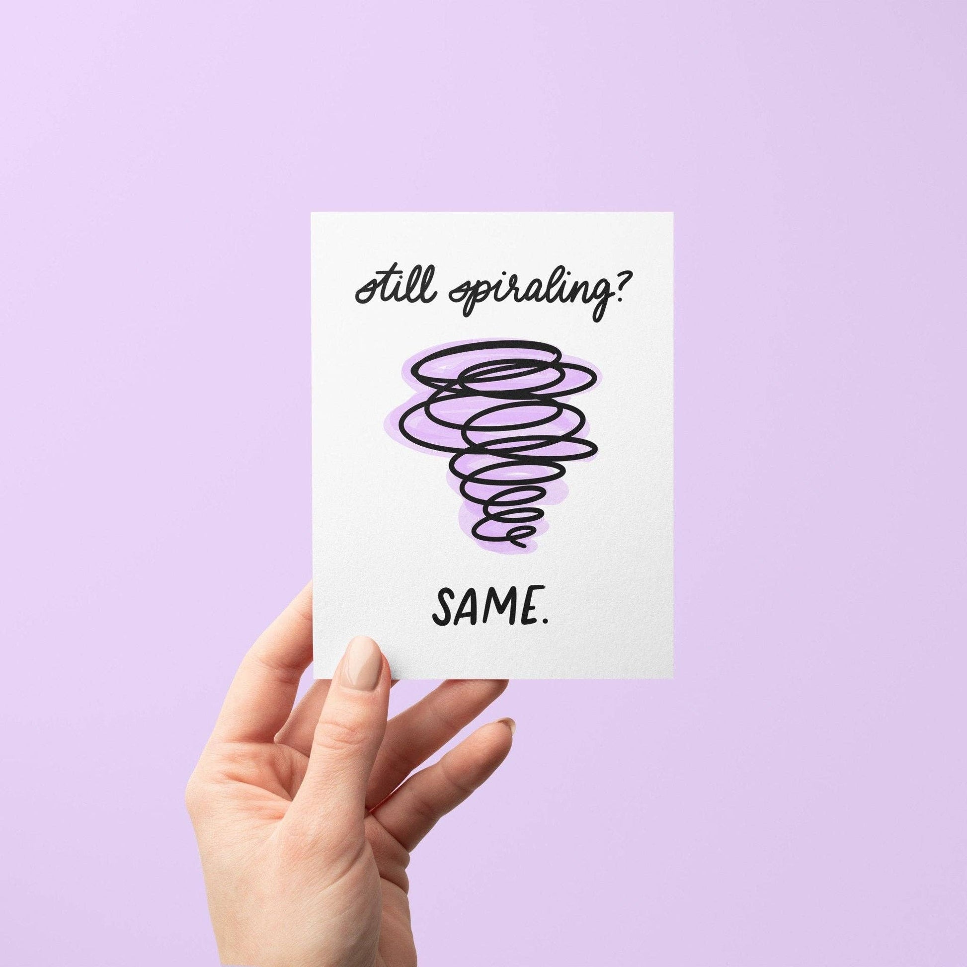 Hand holding a card with 'Still spiraling? Same.' text on a purple background