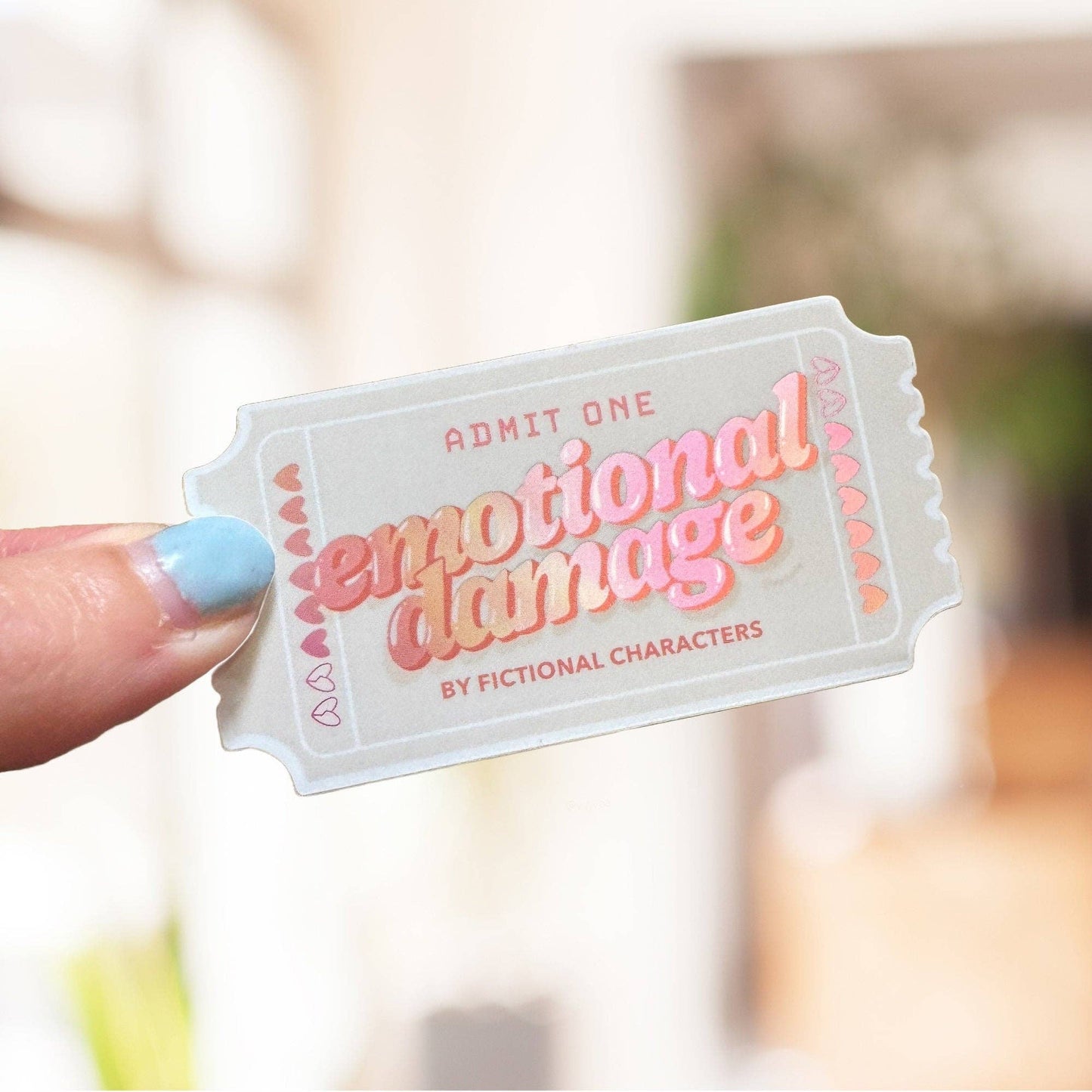 Ticket-shaped card with 'emotional damage' text held by a hand against a blurred background